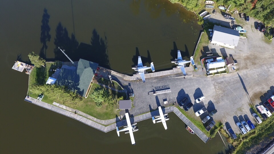 Aerial view of a marina with several seaplanes docked, surrounded by water and greenery. A building with a green roof is visible, along with boats and cars parked nearby.