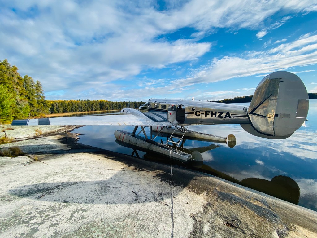 A floatplane parked on a rocky shoreline next to a calm lake, surrounded by trees and under a partly cloudy sky.