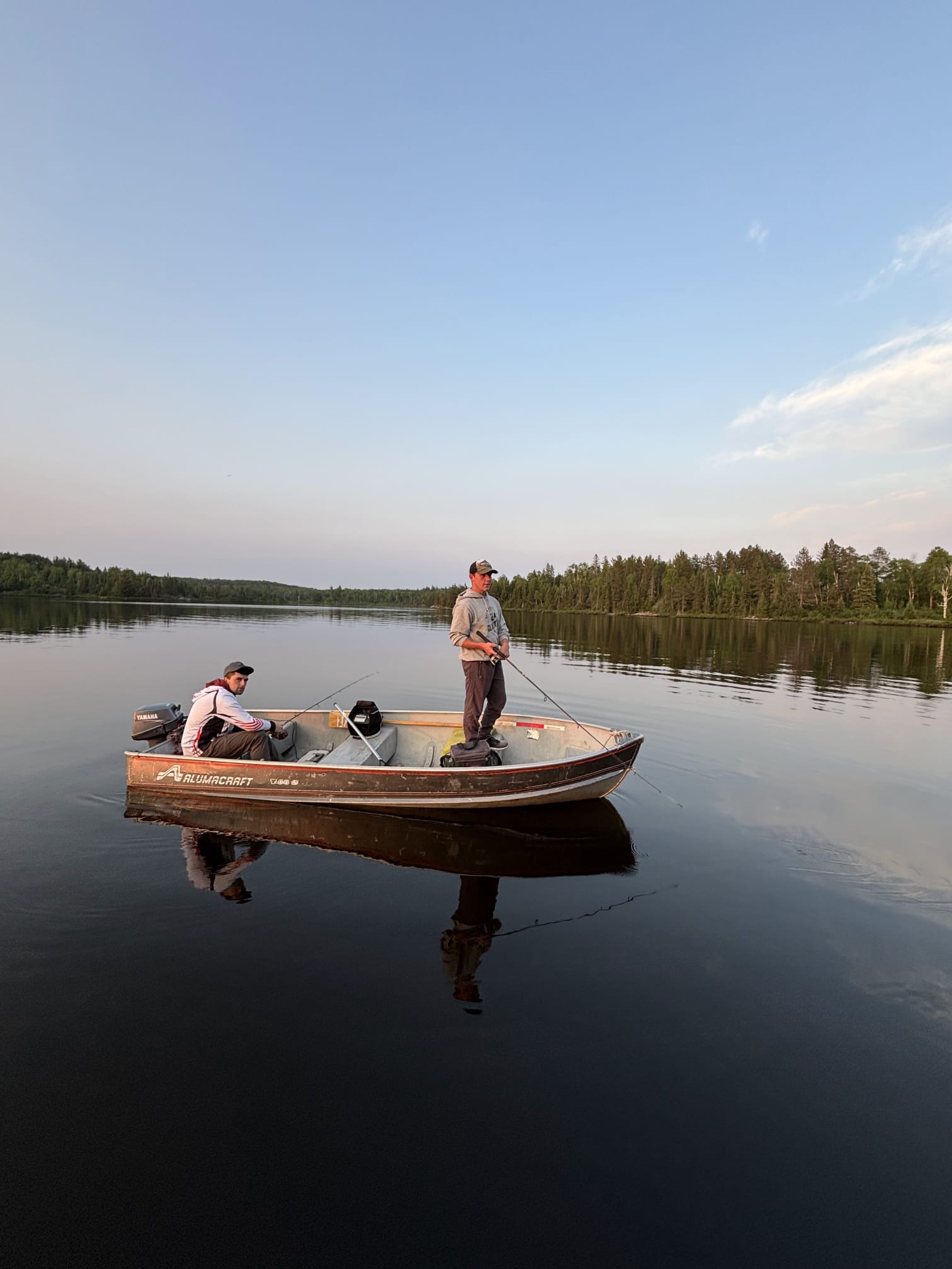 Two men fishing in a small boat on a calm lake surrounded by trees under a clear sky.