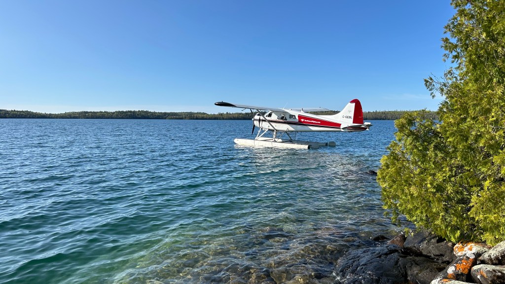 A floatplane parked near a rocky shoreline on a clear lake with a blue sky and trees in the background.