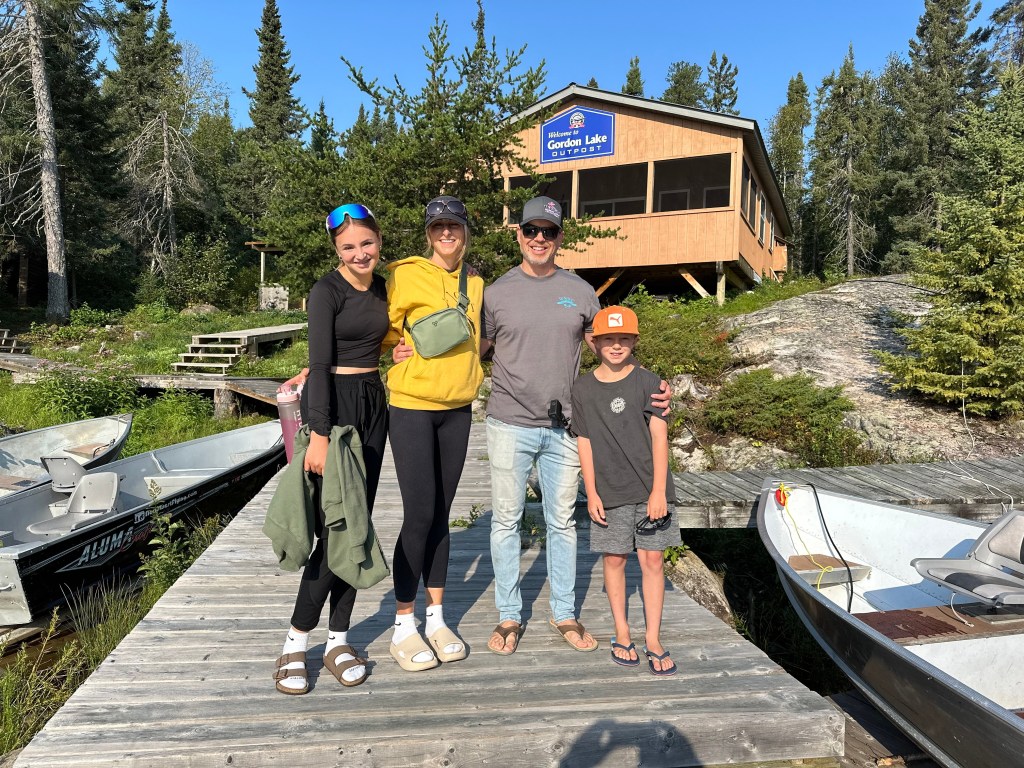 A family standing on a wooden dock by a lake, with a cabin in the background surrounded by trees. The group consists of four people: two women, one man, and a boy, all enjoying a sunny day.