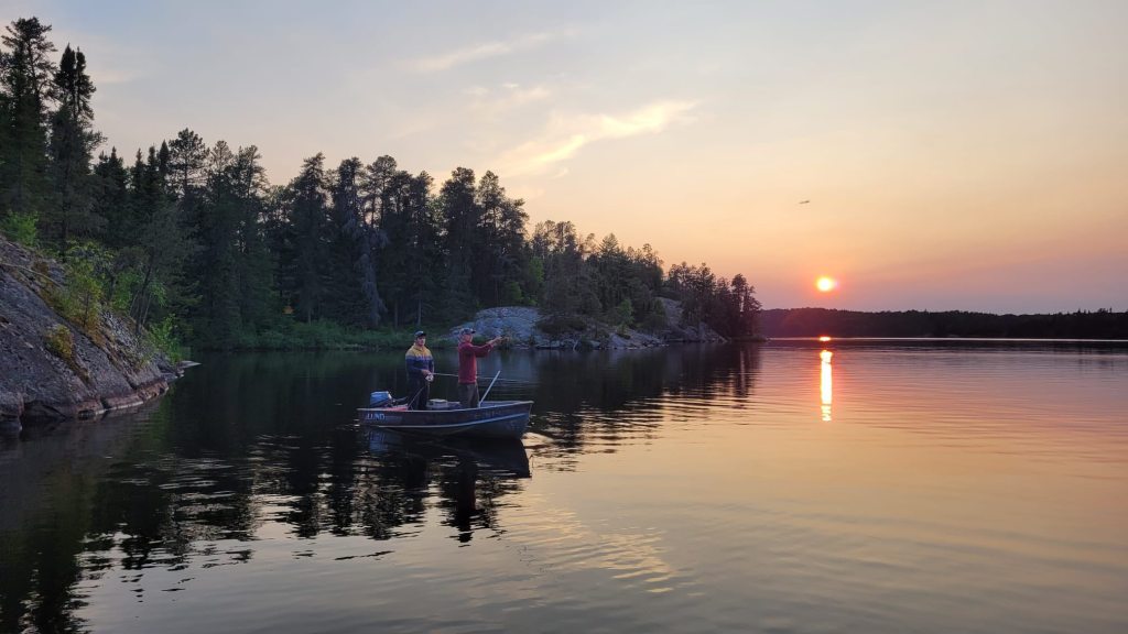 Two fishermen on a small boat in a calm lake at sunset, surrounded by dense forest and rocky shoreline.
