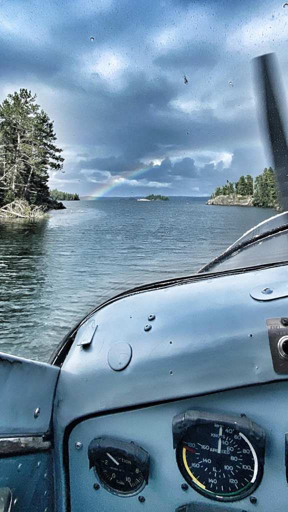 View from the cockpit of a small aircraft flying over a lake, showing water, islands, trees, and a rainbow in the cloudy sky.