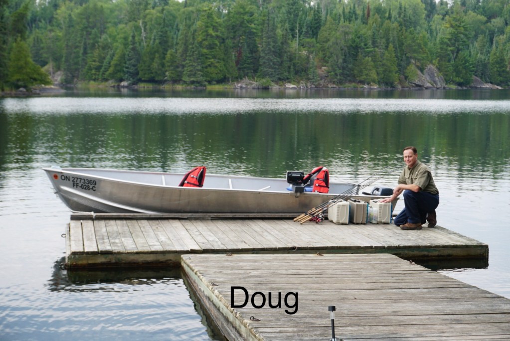 A man kneels on a wooden dock by a tranquil lake, next to a silver boat with an outboard motor and fishing gear.