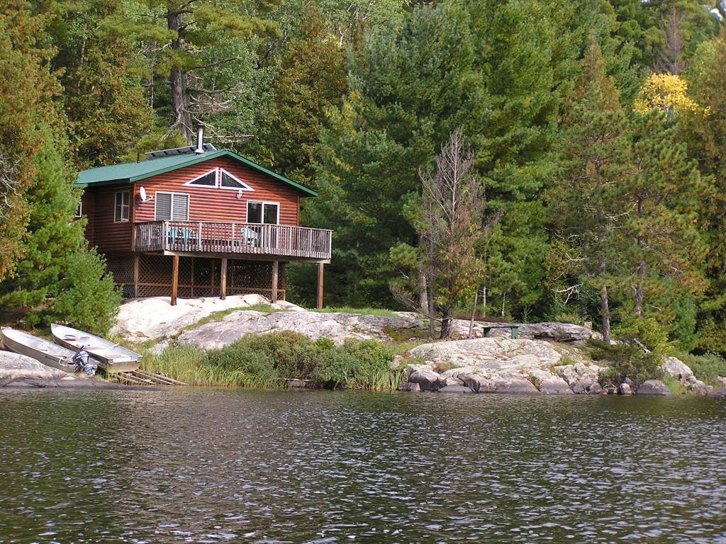 A rustic cabin with a green roof, situated on rocky terrain by a calm lake, surrounded by lush trees.