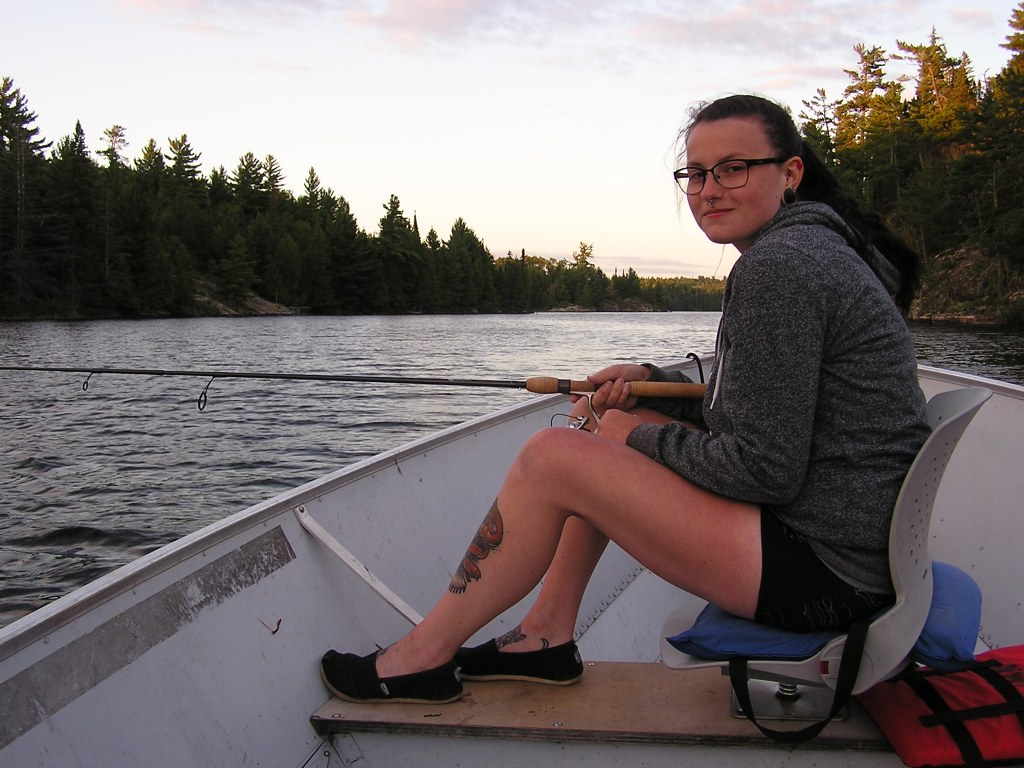 A young woman in glasses sitting in a small boat, fishing on a serene lake surrounded by trees during sunset.