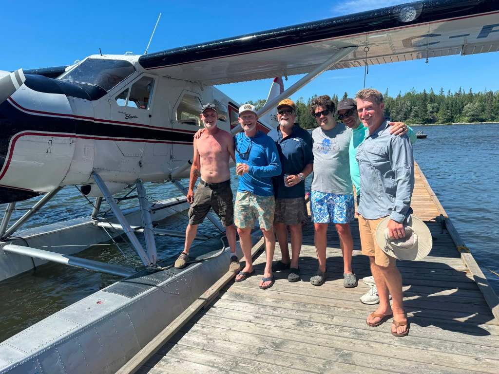 Six longtime friends together on the dock in front of a Northwest Flying Beaver floatplane, Nestor Falls, Ontario, July 2026